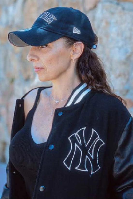 Slide 3:woman wearing black new york yankees bomber jacket and a blue cap